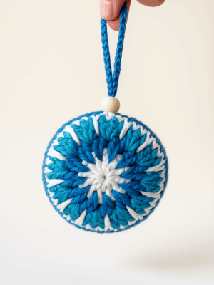 Adorno navideño artesanal en crochet con forma de estrella en tonos azules y blanco, colgante con cuenta de madera sobre fondo claro.