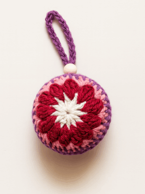 Adorno navideño artesanal de crochet en tonos rojos, rosas y blanco con detalle de cuenta de madera sobre fondo blanco.