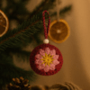 Bola Flor Rosa en árbol de Navidad natural Bola de Navidad de crochet colgada en un árbol real con luces cálidas, en un ambiente acogedor con chimenea.