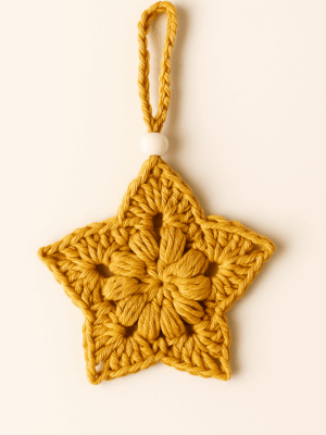 Estrella de Navidad tejida a mano en hilo de algodón natural, con cuenta de madera y colgador trenzado, en color mostaza.