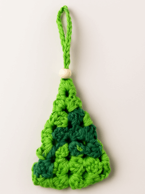 Adorno artesanal en forma de árbol de Navidad tejido a crochet con granny square en tonos verdes y detalle de bolita de madera, sobre fondo claro.